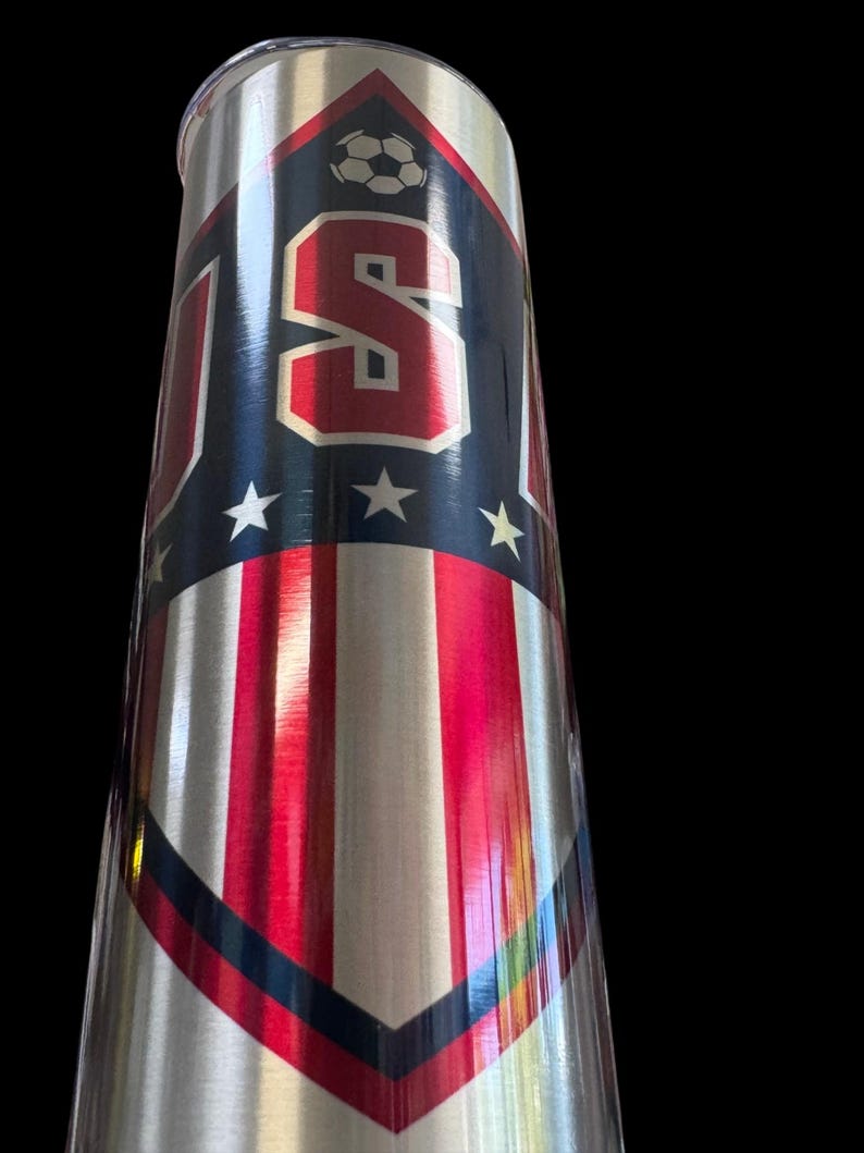Puede incluir: Un vaso de acero inoxidable con un dise&ntilde;o patri&oacute;tico. El vaso presenta las letras "USA" en rojo, blanco y azul, junto con un gr&aacute;fico de bal&oacute;n de f&uacute;tbol y estrellas. El dise&ntilde;o incorpora rayas rojas y blancas.