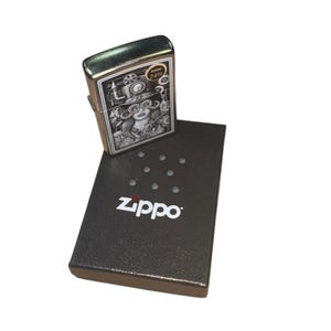 Puede incluir: Un encendedor Zippo plateado con una detallada ilustraci&oacute;n de estilo steampunk. El dise&ntilde;o presenta a una mujer con gafas y engranajes. El encendedor est&aacute; sobre una caja negra Zippo con el nombre de la marca en blanco.
