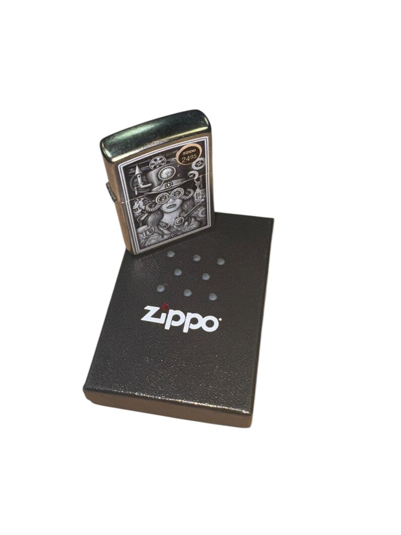 Puede incluir: Un encendedor Zippo plateado con una detallada ilustraci&oacute;n de estilo steampunk. El dise&ntilde;o presenta a una mujer con gafas y engranajes. El encendedor est&aacute; sobre una caja negra Zippo con el nombre de la marca en blanco.