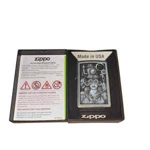 Puede incluir: Un encendedor Zippo a prueba de viento en una caja negra. El encendedor plateado presenta una ilustraci&oacute;n de estilo steampunk de una mujer con gafas y engranajes. La caja dice "Made in USA" e incluye advertencias de seguridad.