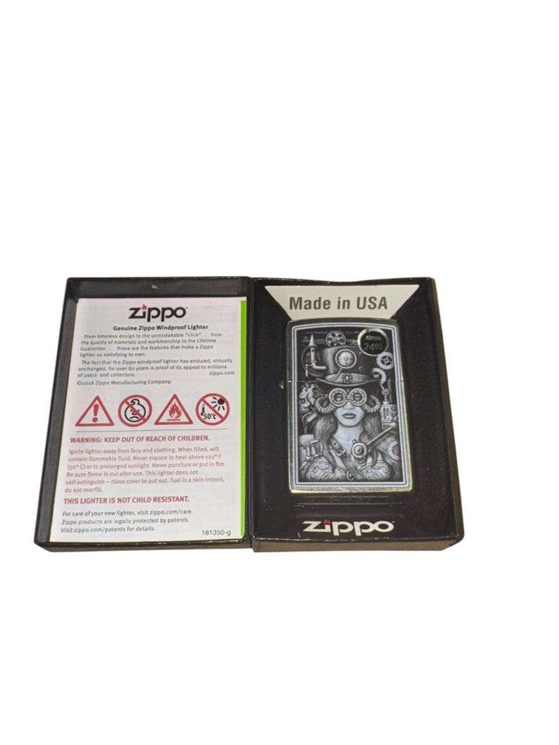 Puede incluir: Un encendedor Zippo a prueba de viento en una caja negra. El encendedor plateado presenta una ilustraci&oacute;n de estilo steampunk de una mujer con gafas y engranajes. La caja dice "Made in USA" e incluye advertencias de seguridad.