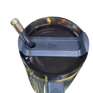 May include: A close-up view of a black tumbler with a gray lid and a clear straw. The tumbler has a gold band near the top and a gray handle. The lid features a small embossed logo. The straw is inserted into the lid.