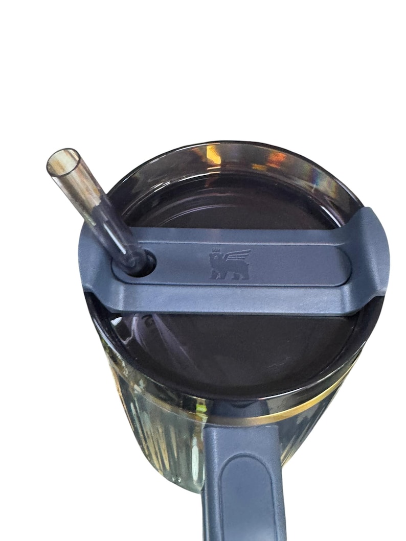 May include: A close-up view of a black tumbler with a gray lid and a clear straw. The tumbler has a gold band near the top and a gray handle. The lid features a small embossed logo. The straw is inserted into the lid.