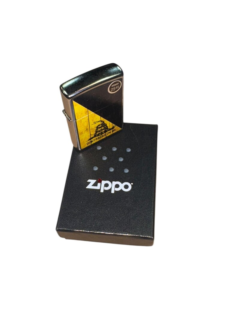 Puede incluir: Un encendedor Zippo con un dise&ntilde;o negro y amarillo con la frase "DON'T TREAD ON ME". El encendedor est&aacute; encima de su caja negra, que tiene el logotipo de Zippo impreso.