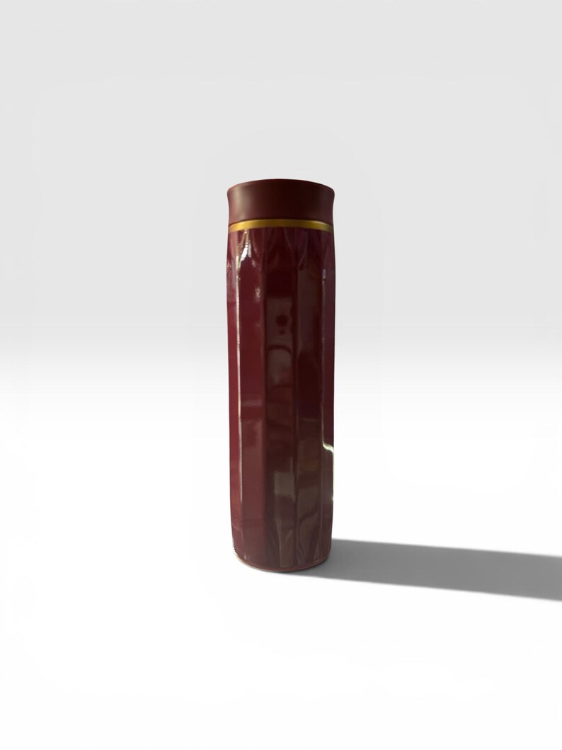 May include: A tall, maroon-colored travel mug with a gold band near the top. The mug has a ridged design and a dark brown lid. The mug is standing upright on a white surface.