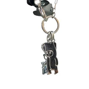 May include: A silver-colored keychain featuring a cartoon figure with a hat and a small animal. The keychain is attached to a black fabric piece and a silver chain. The figure is holding a small object.