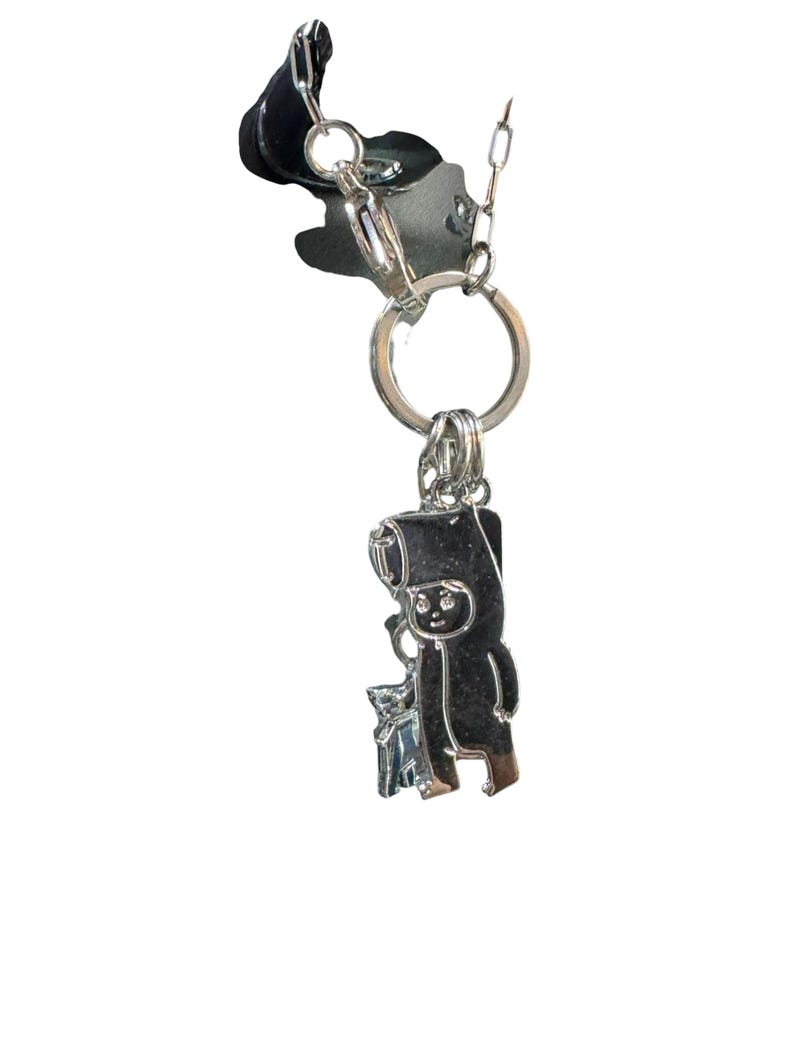 May include: A silver-colored keychain featuring a cartoon figure with a hat and a small animal. The keychain is attached to a black fabric piece and a silver chain. The figure is holding a small object.