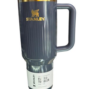 May include: A gray Stanley Fluted Tumbler with a black lid and handle. The tumbler has a gold accent and the Stanley logo in gold. The label reads "The Quencher H2.0 Fluted Tumbler" and "All Day Hydration".