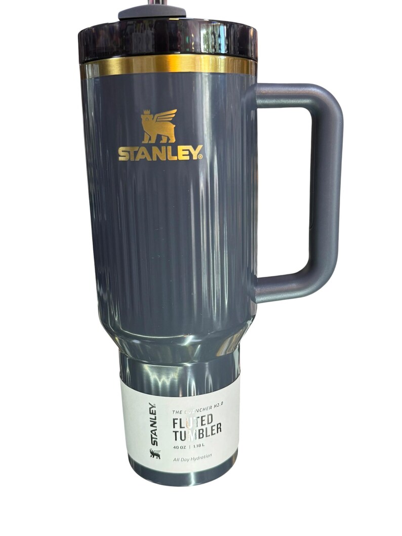 May include: A gray Stanley Fluted Tumbler with a black lid and handle. The tumbler has a gold accent and the Stanley logo in gold. The label reads "The Quencher H2.0 Fluted Tumbler" and "All Day Hydration".