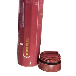 May include: A maroon Stanley thermos with a gold rim and a matching maroon lid. The thermos has a cylindrical shape with a ridged design. The Stanley logo is printed on the side.
