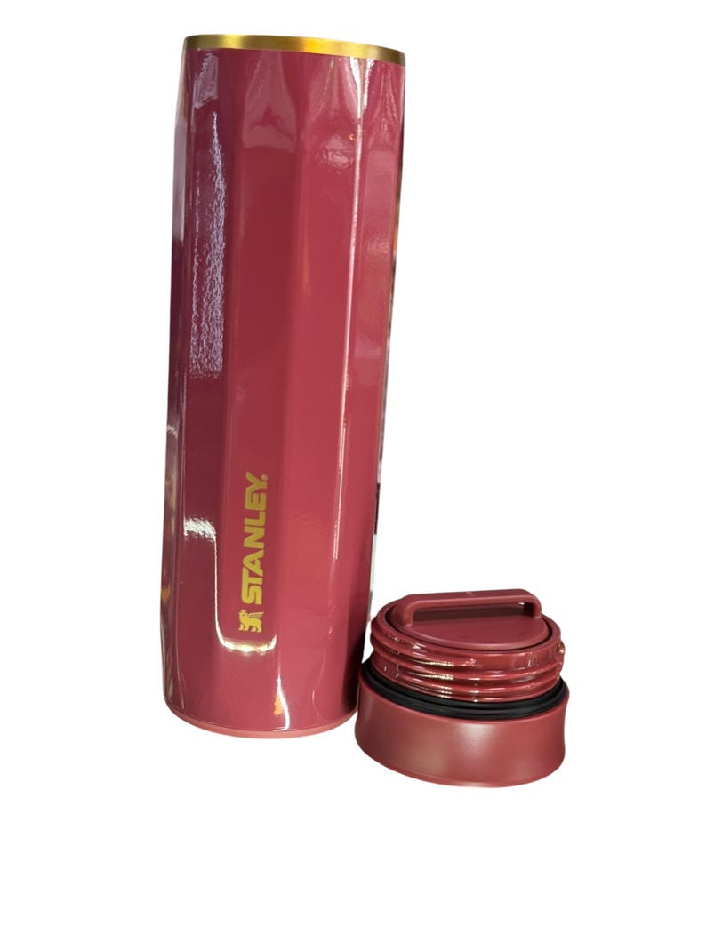May include: A maroon Stanley thermos with a gold rim and a matching maroon lid. The thermos has a cylindrical shape with a ridged design. The Stanley logo is printed on the side.