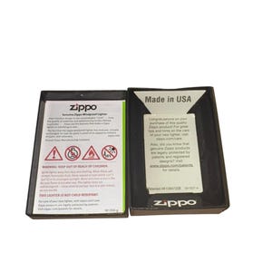 Puede incluir: Dos cajas negras de encendedores Zippo, una abierta y otra cerrada. La caja abierta muestra texto, incluidas advertencias y el logotipo de Zippo. La caja cerrada tiene el texto "Made in USA" y el logotipo de Zippo. Las cajas son rectangulares.