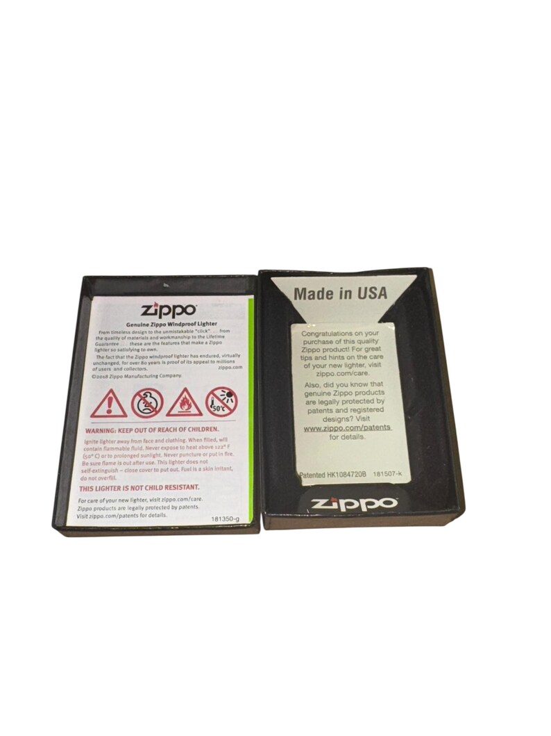 Puede incluir: Dos cajas negras de encendedores Zippo, una abierta y otra cerrada. La caja abierta muestra texto, incluidas advertencias y el logotipo de Zippo. La caja cerrada tiene el texto "Made in USA" y el logotipo de Zippo. Las cajas son rectangulares.