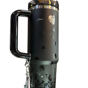 May include: Black travel mug with a silver handle and lid. The mug features a floral design and the name "Jennie" in cursive. A silver charm hangs from the handle. The mug is designed to keep beverages hot or cold.