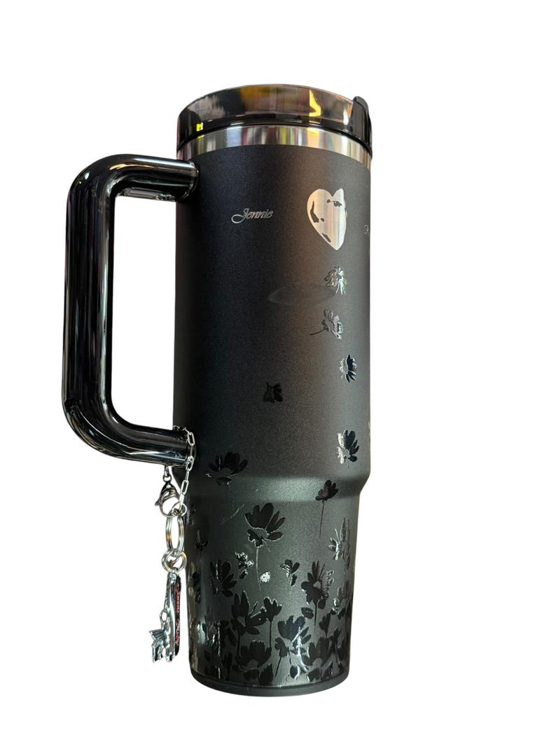 May include: Black travel mug with a silver handle and lid. The mug features a floral design and the name "Jennie" in cursive. A silver charm hangs from the handle. The mug is designed to keep beverages hot or cold.