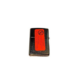 Puede incluir: Un encendedor de metal plateado con forma rectangular. Una etiqueta de advertencia roja est&aacute; adherida, mostrando un s&iacute;mbolo de prohibido fumar e instrucciones de seguridad. El encendedor est&aacute; cerrado y parece sin usar.