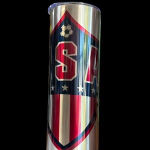 Puede incluir: Un vaso de acero inoxidable con un dise&ntilde;o patri&oacute;tico. El vaso presenta un escudo con las letras "USA" en rayas rojas y blancas, detalles azules y un gr&aacute;fico de bal&oacute;n de f&uacute;tbol. El dise&ntilde;o est&aacute; sobre un fondo de metal cepillado.