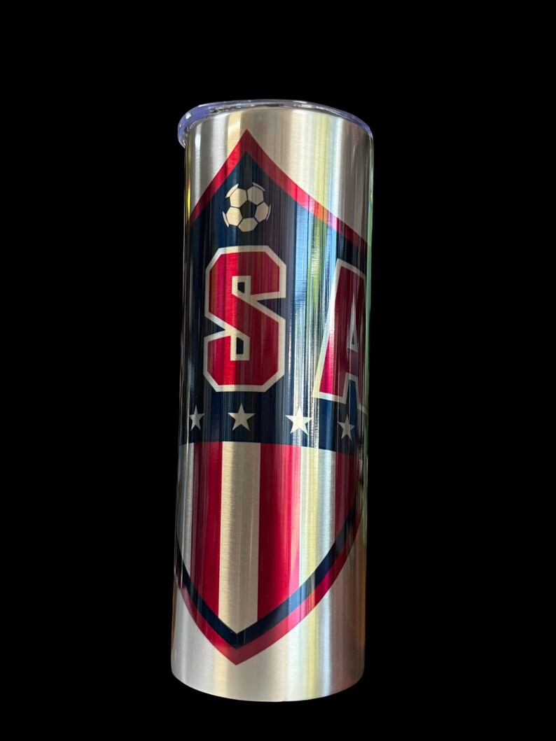 Puede incluir: Un vaso de acero inoxidable con un dise&ntilde;o patri&oacute;tico. El vaso presenta un escudo con las letras "USA" en rayas rojas y blancas, detalles azules y un gr&aacute;fico de bal&oacute;n de f&uacute;tbol. El dise&ntilde;o est&aacute; sobre un fondo de metal cepillado.
