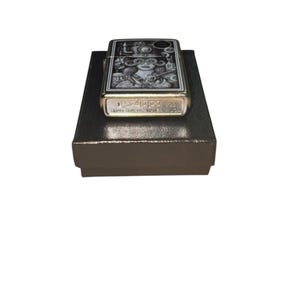 Puede incluir: Un encendedor Zippo con una ilustraci&oacute;n detallada en blanco y negro sobre una superficie met&aacute;lica dorada. El dise&ntilde;o presenta una mujer con gafas y otros elementos de inspiraci&oacute;n steampunk. El encendedor est&aacute; sobre una caja marr&oacute;n oscuro.