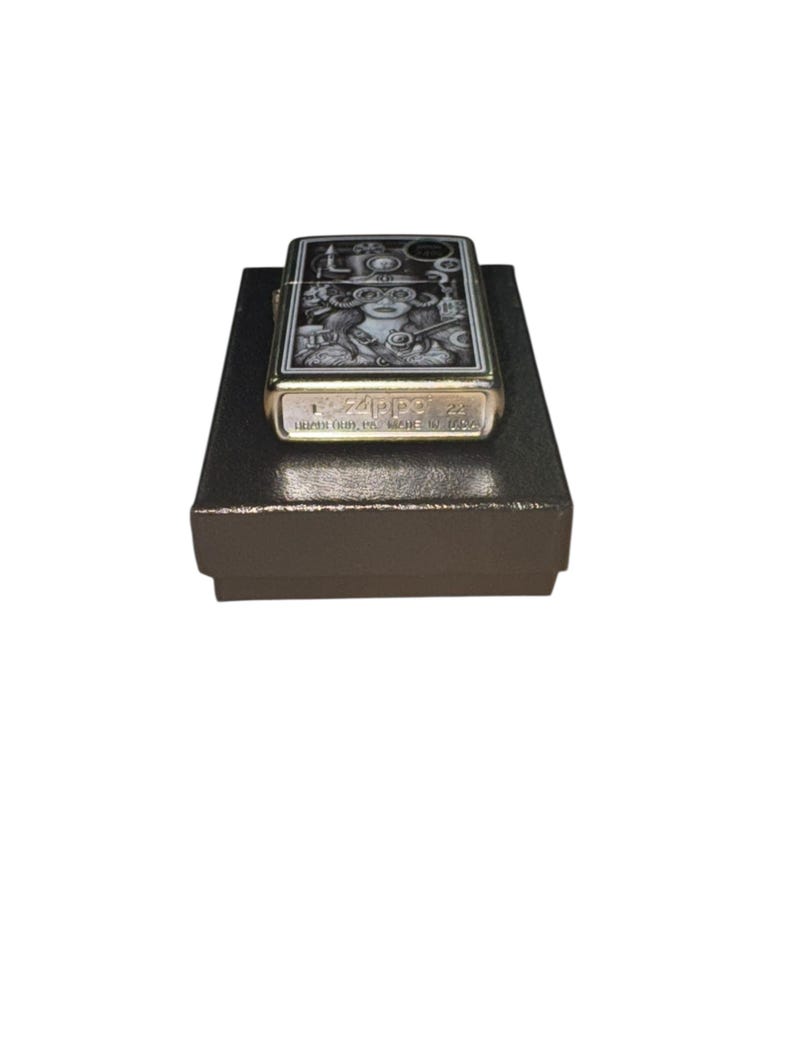 Puede incluir: Un encendedor Zippo con una ilustraci&oacute;n detallada en blanco y negro sobre una superficie met&aacute;lica dorada. El dise&ntilde;o presenta una mujer con gafas y otros elementos de inspiraci&oacute;n steampunk. El encendedor est&aacute; sobre una caja marr&oacute;n oscuro.