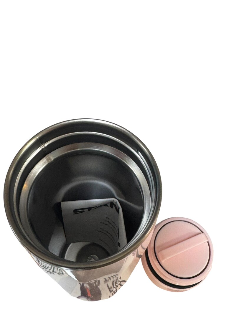 Peut inclure: Un gobelet isotherme en acier inoxydable avec un couvercle rose clair et un joint noir. Le gobelet pr&eacute;sente un motif avec du texte et des images. L'int&eacute;rieur est argent&eacute;, et un papier pli&eacute; est &agrave; l'int&eacute;rieur.