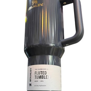 May include: A gray Stanley Fluted Tumbler with a black handle. The tumbler has a gold rim and the Stanley logo. The label reads "THE QUENCHER H2.0 FLUTED TUMBLER 40 OZ | 1.18 L All Day Hydration."