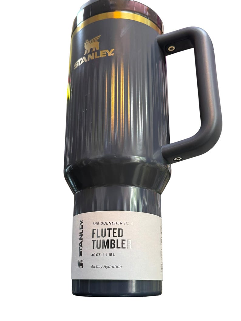 May include: A gray Stanley Fluted Tumbler with a black handle. The tumbler has a gold rim and the Stanley logo. The label reads "THE QUENCHER H2.0 FLUTED TUMBLER 40 OZ | 1.18 L All Day Hydration."