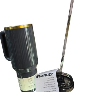 May include: A black Stanley travel mug with a gold rim and handle, next to a clear straw and lid. A white paper with "STANLEY" and care instructions is also present.