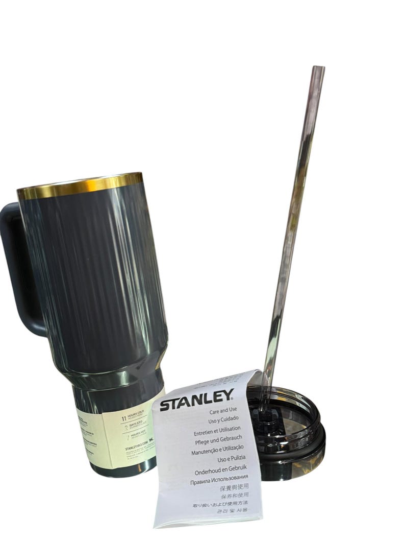 May include: A black Stanley travel mug with a gold rim and handle, next to a clear straw and lid. A white paper with "STANLEY" and care instructions is also present.
