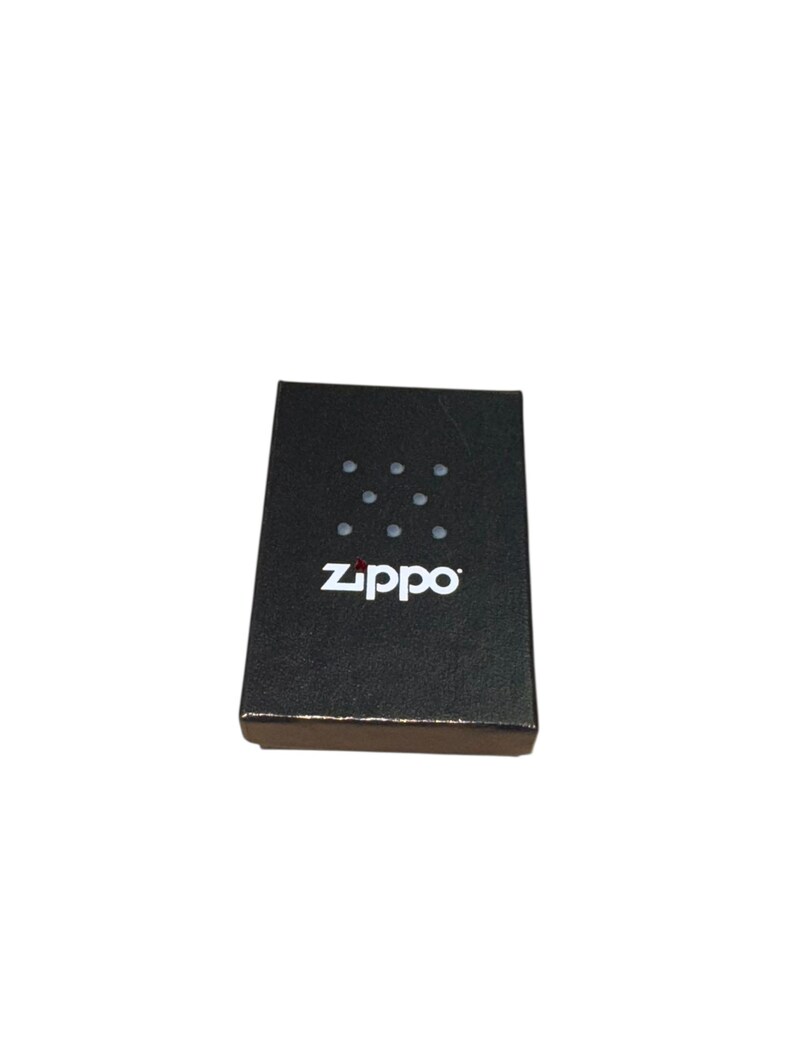Puede incluir: Una caja rectangular negra con la marca "Zippo" en blanco. La caja tiene un patr&oacute;n de puntos en la parte superior. La caja es probablemente para un encendedor o un producto similar.