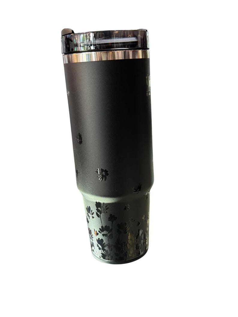 May include: A black insulated tumbler with a clear lid. The tumbler features a floral design at the bottom and small floral accents. The tumbler has a silver rim and a black handle. The tumbler is designed to keep beverages hot or cold.