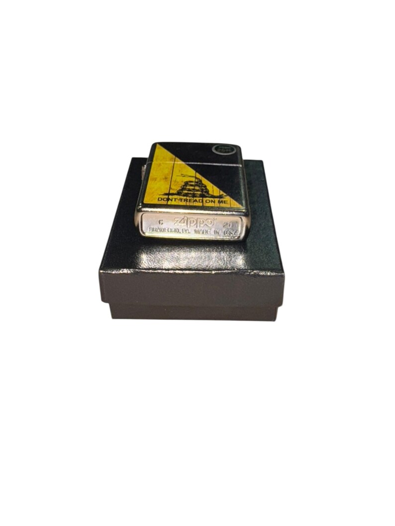Puede incluir: Un encendedor Zippo plateado con un dise&ntilde;o negro y amarillo con la frase "DON'T TREAD ON ME". El encendedor est&aacute; sobre una caja negra. El encendedor tiene el logotipo de Zippo y el texto "BRADFORD, PA. MADE IN USA."