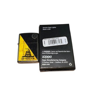 Puede incluir: Un encendedor Zippo negro y amarillo con el texto "DONT TREAD ON ME" y un dise&ntilde;o de serpiente. El encendedor est&aacute; junto a su caja negra, que incluye el texto "Genuine Zippo Lighter Made in USA".