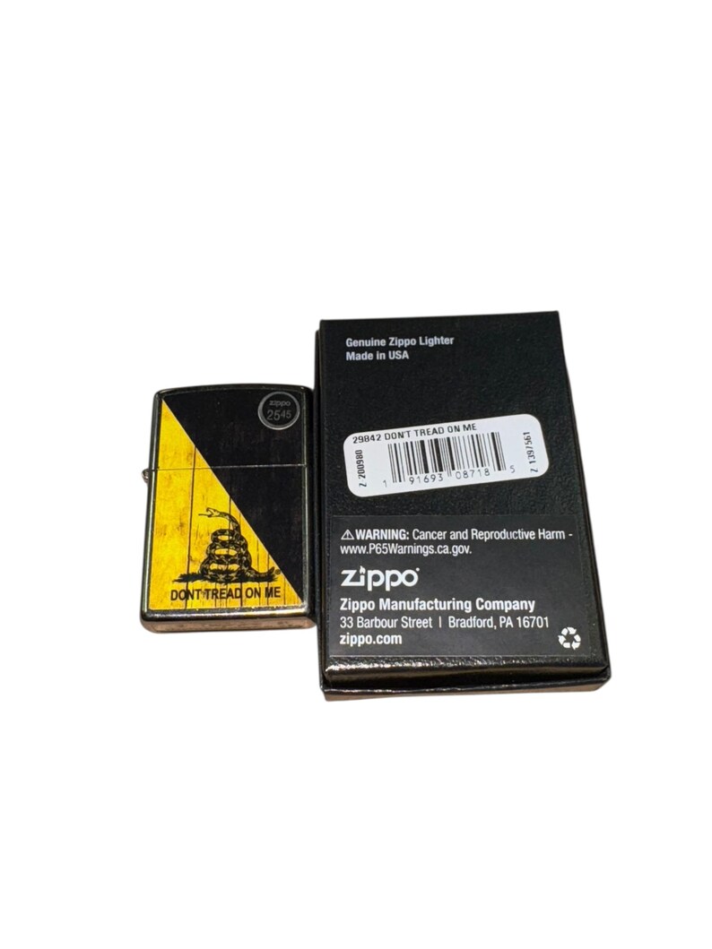 Puede incluir: Un encendedor Zippo negro y amarillo con el texto "DONT TREAD ON ME" y un dise&ntilde;o de serpiente. El encendedor est&aacute; junto a su caja negra, que incluye el texto "Genuine Zippo Lighter Made in USA".