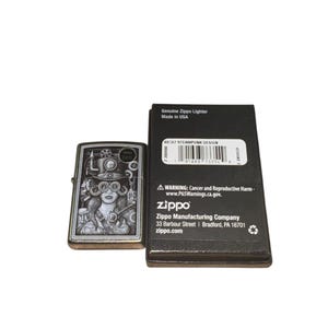Puede incluir: Un encendedor Zippo plateado con un dise&ntilde;o steampunk que presenta a una mujer con gafas y engranajes. El encendedor est&aacute; junto a su caja negra, que incluye el texto "Genuine Zippo Lighter Made in USA".
