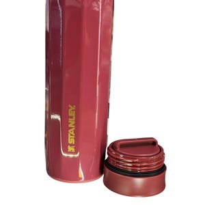 May include: A maroon Stanley thermos with a gold rim and a matching maroon lid. The thermos has a geometric design and the Stanley logo is visible on the side. The lid is off to the side.