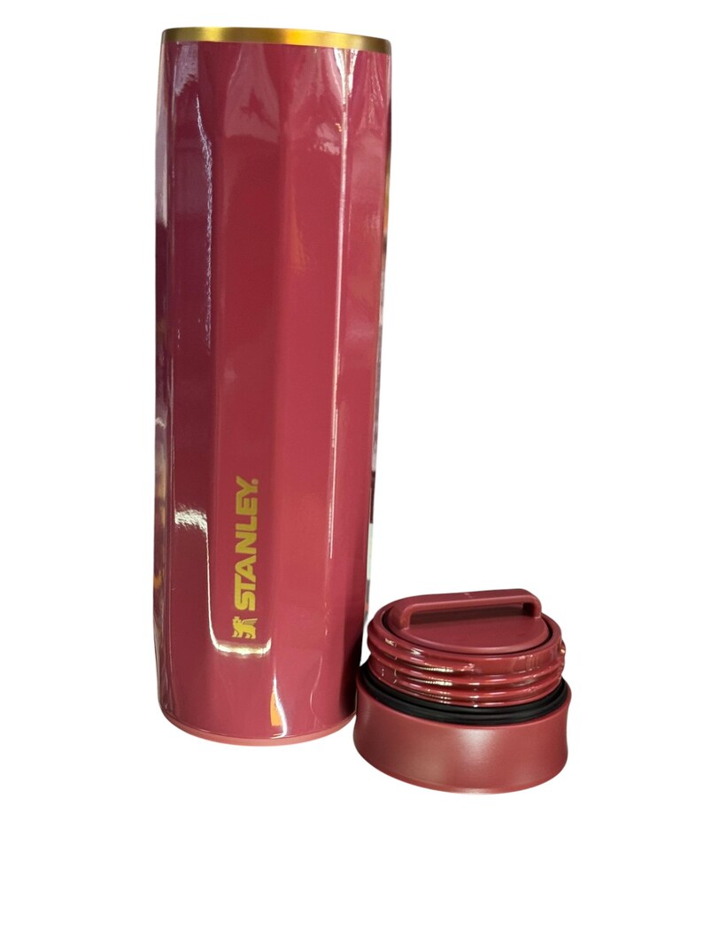 May include: A maroon Stanley thermos with a gold rim and a matching maroon lid. The thermos has a geometric design and the Stanley logo is visible on the side. The lid is off to the side.