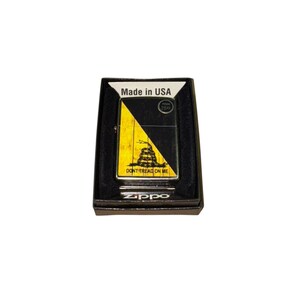 Puede incluir: Un encendedor Zippo negro en su caja original. El encendedor presenta un dise&ntilde;o amarillo y negro con las palabras "DON'T TREAD ON ME" y una serpiente enroscada. La caja tiene la inscripci&oacute;n "Made in USA".
