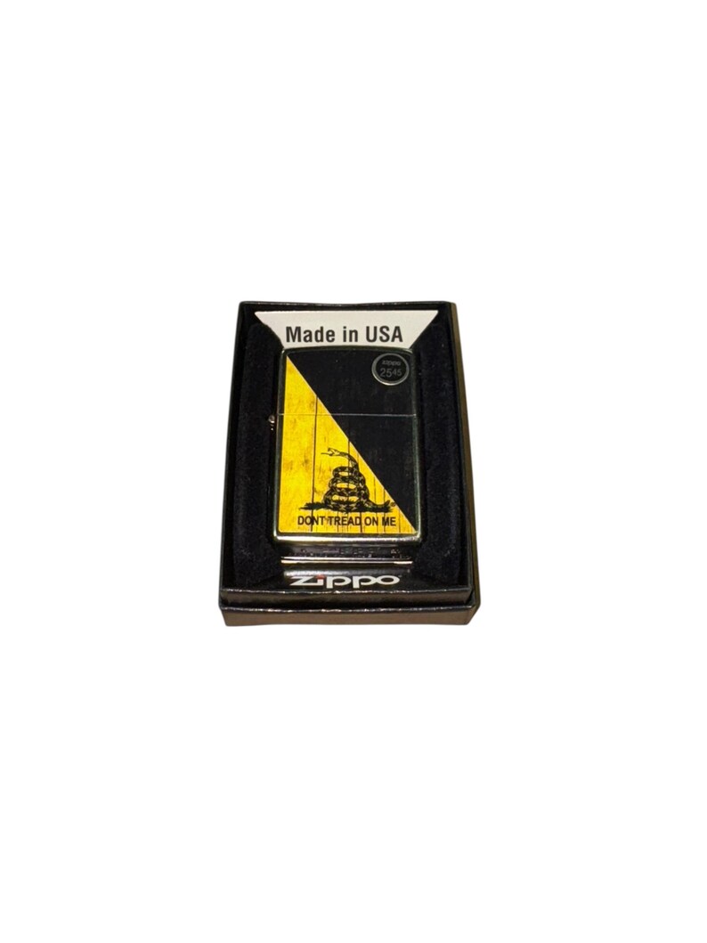 Puede incluir: Un encendedor Zippo negro en su caja original. El encendedor presenta un dise&ntilde;o amarillo y negro con las palabras "DON'T TREAD ON ME" y una serpiente enroscada. La caja tiene la inscripci&oacute;n "Made in USA".