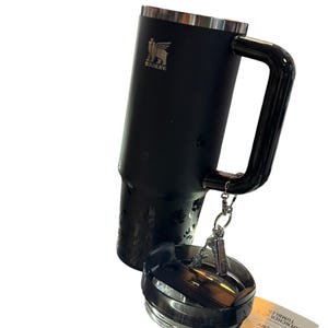 May include: A black Stanley Quencher tumbler with a handle and a lid. The tumbler has a silver rim and the Stanley logo. The lid is detached and has a silver and black design. A tag with the words "QUENCHER H2.0 TUMBLER" is attached.