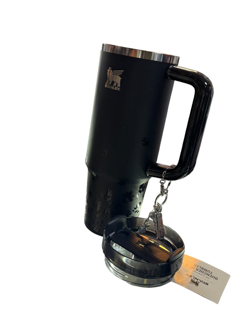 May include: A black Stanley Quencher tumbler with a handle and a lid. The tumbler has a silver rim and the Stanley logo. The lid is detached and has a silver and black design. A tag with the words "QUENCHER H2.0 TUMBLER" is attached.