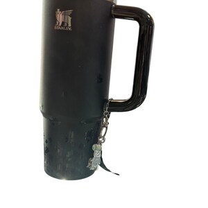 May include: Black Stanley travel mug with a silver rim and handle. The mug has a silver charm attached to the handle. The Stanley logo is visible on the side of the mug. The mug is designed to keep beverages hot or cold.