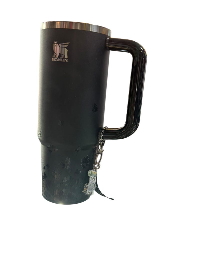 May include: Black Stanley travel mug with a silver rim and handle. The mug has a silver charm attached to the handle. The Stanley logo is visible on the side of the mug. The mug is designed to keep beverages hot or cold.