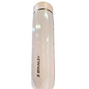 Peut inclure: Une thermos Stanley blanche avec un couvercle rose clair et une bande dor&eacute;e. Le logo Stanley est imprim&eacute; en noir sur le c&ocirc;t&eacute;. La thermos est cylindrique et semble &ecirc;tre en m&eacute;tal.