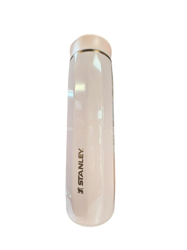 Peut inclure: Une thermos Stanley blanche avec un couvercle rose clair et une bande dor&eacute;e. Le logo Stanley est imprim&eacute; en noir sur le c&ocirc;t&eacute;. La thermos est cylindrique et semble &ecirc;tre en m&eacute;tal.