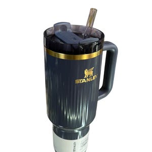 May include: A dark blue Stanley tumbler with a handle and a clear straw. The tumbler has a gold band and the Stanley logo in gold. The lid is dark blue with a sliding closure. The tumbler has a ribbed design.