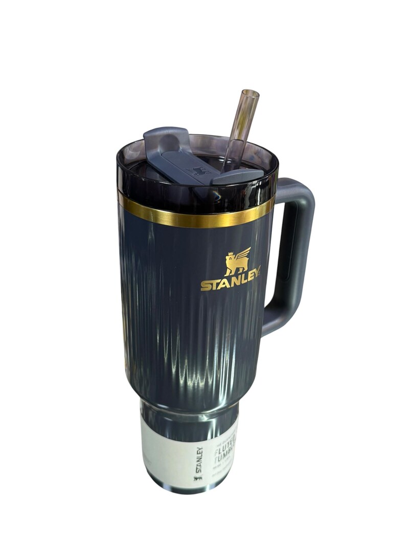 May include: A dark blue Stanley tumbler with a handle and a clear straw. The tumbler has a gold band and the Stanley logo in gold. The lid is dark blue with a sliding closure. The tumbler has a ribbed design.