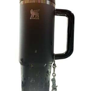 May include: Black Stanley travel mug with a large handle and a floral design. The mug has a black lid and a silver charm of a dog. The Stanley logo is visible on the front.