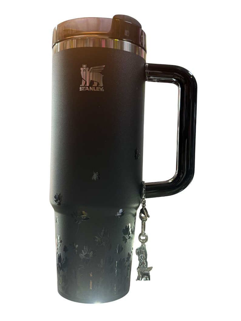 May include: Black Stanley travel mug with a large handle and a floral design. The mug has a black lid and a silver charm of a dog. The Stanley logo is visible on the front.