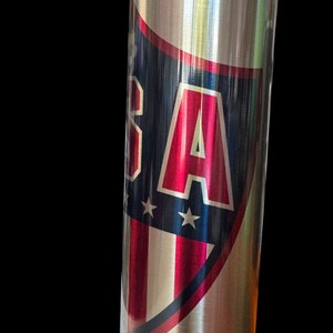 Puede incluir: Un vaso de acero inoxidable con un dise&ntilde;o patri&oacute;tico. El vaso presenta un escudo con las letras "USA" en rojo, blanco y azul, junto con estrellas y rayas. El dise&ntilde;o est&aacute; sobre un fondo plateado met&aacute;lico.